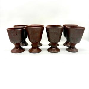 May include: A set of seven brown ceramic goblets with a pedestal base. The goblets have a wide, slightly flared rim and a dark, earthy tone. The stem is short and the base is round.