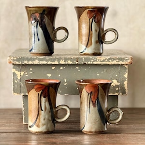 May include: Four ceramic mugs with a unique design. Each mug features a brown rim, a tan and white body, and a dark brown and red floral design. The mugs have a rounded handle and are displayed on a weathered wooden surface.