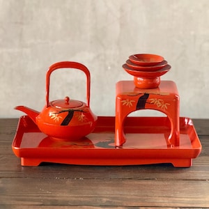 May include: A red lacquered Japanese tea set. The set includes a teapot with a handle, a small table with stacked bowls, and a rectangular tray. The teapot and table are decorated with black and gold floral designs.