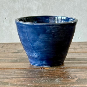 May include: A handmade, deep blue ceramic bowl with a slightly flared shape. The bowl has a smooth, glossy finish and sits on a wooden surface. The interior is a darker shade of blue.