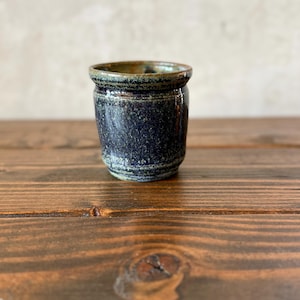 May include: A small, handcrafted ceramic pot with a dark blue speckled glaze. The rim and interior have a greenish-brown hue. The cylindrical shape sits on a wooden surface, showcasing the texture and color variations of the pottery.