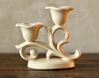 May include: Cream-coloured ceramic candle holder with two tulip-shaped candle cups. The candle holder has a decorative floral design with a curved base and stem. The piece is set on a dark wooden surface.