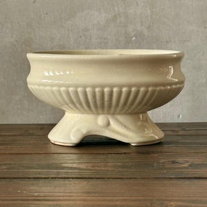 May include: Cream-colored ceramic planter with a pedestal base. The planter has a wide, shallow bowl with a ribbed design around the middle. The base features a decorative leaf design. The planter is suitable for small plants or decorative use.