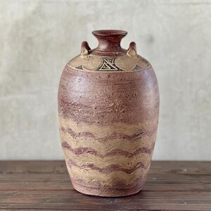 May include: A handcrafted, reddish-brown ceramic vase with a textured surface. The vase features a wavy, tan pattern around the body and decorative elements near the top, including a geometric design and small handles. The vase is displayed on a wooden surface.
