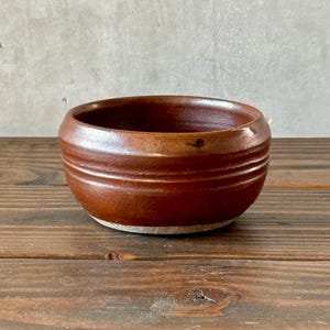 May include: A small, round, brown ceramic bowl with a textured exterior. The bowl has a wide opening and a slightly raised base. The exterior features horizontal grooves. The bowl is set on a wooden surface.