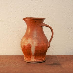 May include: A ceramic pitcher with a rounded body and a curved handle. The exterior features a speckled terracotta orange glaze with a prominent white drip pattern running down the center of the vessel, creating a textured appearance.