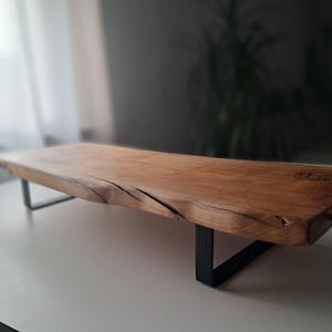 Pode incluir: Uma bandeja de servir de madeira longa com um acabamento natural e rústico. A bandeja tem uma borda viva e é sustentada por duas pernas de metal preto. A bandeja tem uma alça de metal preto em uma extremidade.