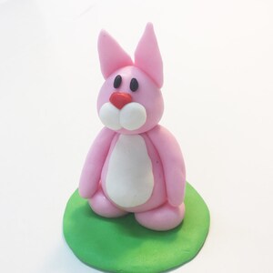 May include: A pink fondant bunny rabbit with white accents and black eyes, sitting on a green fondant base.