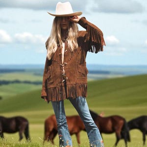 Pode incluir: Casaco de camurça castanho com franjas, chapéu de cowboy creme e calças de ganga azuis com bordados florais. O casaco tem franjas nas mangas e na bainha. Cavalos pastam ao fundo numa colina gramada.