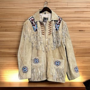 Peut inclure: Veste en daim beige avec franges et détails perlés. La veste présente des broderies de perles décoratives sur le col, les poches et les manches. La veste est boutonnée et de style western classique.