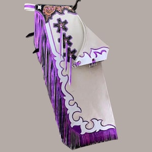 May include: A white and tan leather chaps with purple fringe and decorative accents. The chaps feature a white decorative trim, star-shaped cutouts, and a buckle closure. The top portion has a floral design with brown and gold accents.