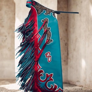 May include: A turquoise leather chaps with red and teal fringe. The chaps feature red decorative accents and a gold-colored design. The initials "DC" are visible on the lower portion of the chaps.