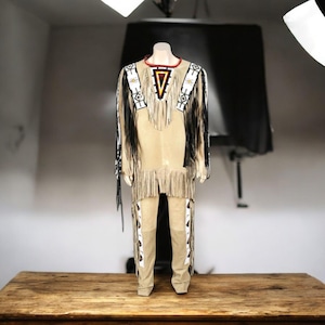 May include: A tan suede-like two-piece outfit with fringe and decorative beading. The top has a red, black, and yellow triangle design on the chest. The pants have decorative white and black patterns on the sides. The outfit is displayed on a mannequin.