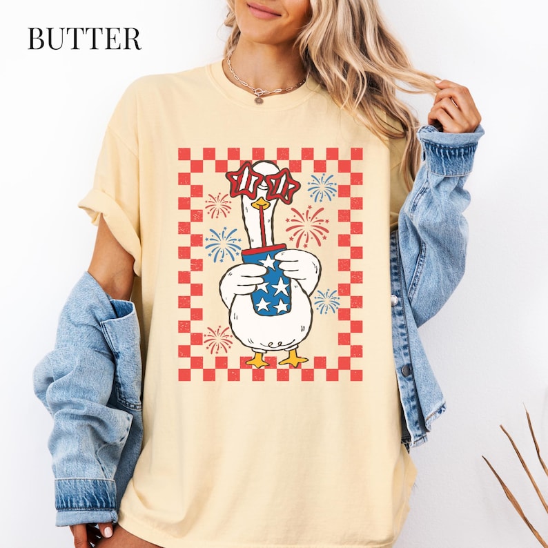 May include: Butter-colored t-shirt featuring a patriotic goose wearing star-shaped glasses and holding a container with stars and stripes. The goose is set against a red and white checkered background with fireworks.