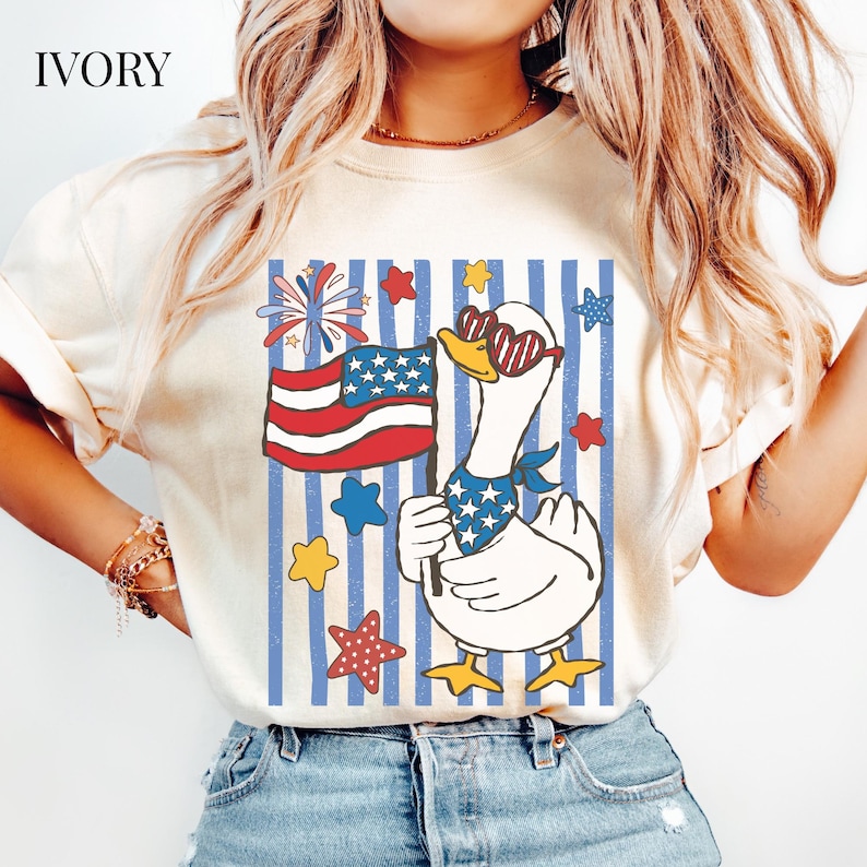 Pu&ograve; includere: T-shirt color avorio con un'oca dei cartoni animati con occhiali da sole a forma di cuore e una bandana patriottica. L'oca tiene in mano una bandiera americana su uno sfondo a strisce blu e bianche, con stelle e fuochi d'artificio.