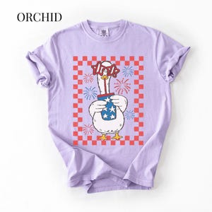 May include: A lilac-colored t-shirt featuring a cartoon goose wearing star-shaped glasses and holding a container with stars. The goose is set against a red and white checkered background with fireworks. The word "ORCHID" is at the top.