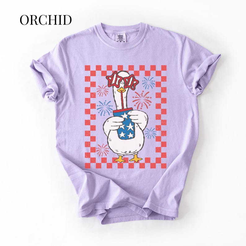 May include: A lilac-colored t-shirt featuring a cartoon goose wearing star-shaped glasses and holding a container with stars. The goose is set against a red and white checkered background with fireworks. The word "ORCHID" is at the top.
