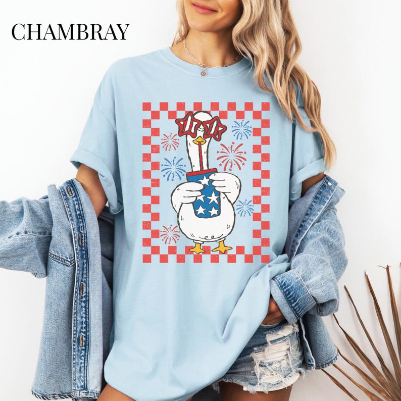 May include: Light blue t-shirt with a graphic of a goose wearing star-shaped glasses and holding a container with stars. The design is set against a red and white checkered background with fireworks. The word "CHAMBRAY" is at the top.