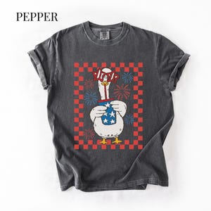 May include: Dark gray t-shirt with a graphic of a goose wearing star-shaped glasses and holding a blue cup with stars. The goose is set against a red and white checkered background with fireworks. The word "PEPPER" is at the top.
