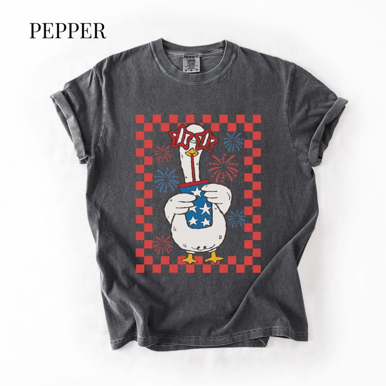 May include: Dark gray t-shirt with a graphic of a goose wearing star-shaped glasses and holding a blue cup with stars. The goose is set against a red and white checkered background with fireworks. The word "PEPPER" is at the top.