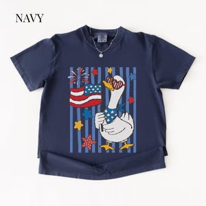 Pu&ograve; includere: T-shirt blu navy con un'oca dei cartoni animati con occhiali da sole e una bandana patriottica, che tiene una bandiera americana. Il design include stelle, strisce e fuochi d'artificio rossi, bianchi e blu. La parola "NAVY" &egrave; in alto a sinistra.