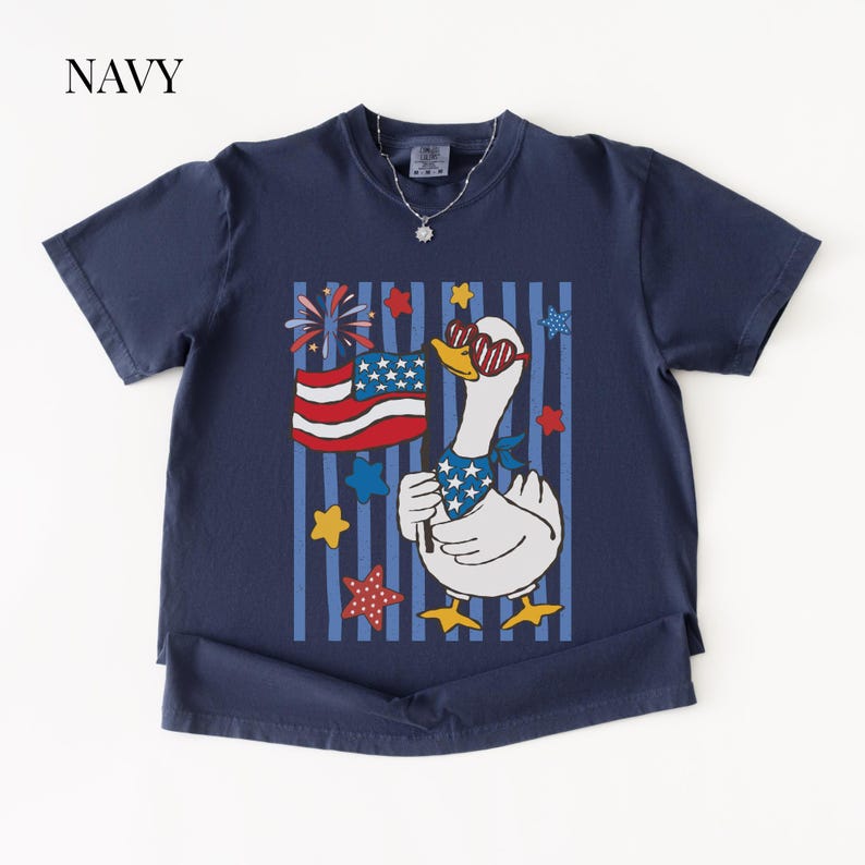 Pu&ograve; includere: T-shirt blu navy con un'oca dei cartoni animati con occhiali da sole e una bandana patriottica, che tiene una bandiera americana. Il design include stelle, strisce e fuochi d'artificio rossi, bianchi e blu. La parola "NAVY" &egrave; in alto a sinistra.