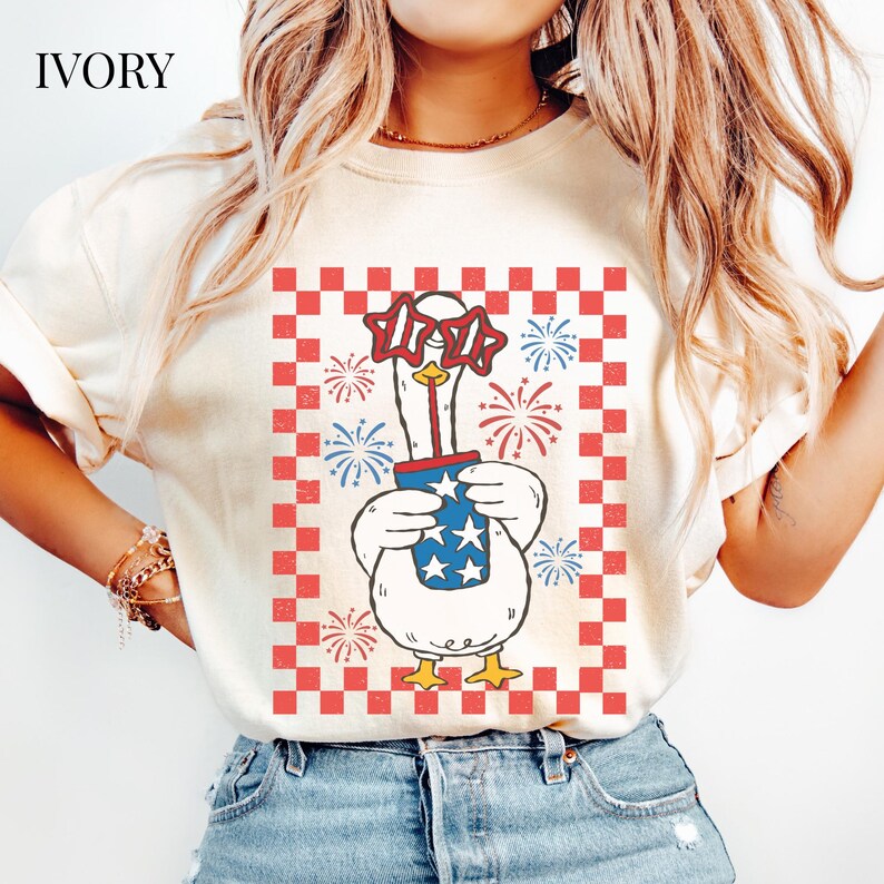 May include: Ivory-colored t-shirt featuring a cartoon goose wearing star-shaped sunglasses and holding a blue cup with stars. The design includes fireworks and a red and white checkered border, perfect for patriotic celebrations.