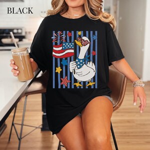 Pu&ograve; includere: T-shirt nero con un'oca dei cartoni animati con occhiali da sole e una bandana patriottica, che tiene una bandiera americana. Il design include stelle e strisce rosse, bianche e blu.