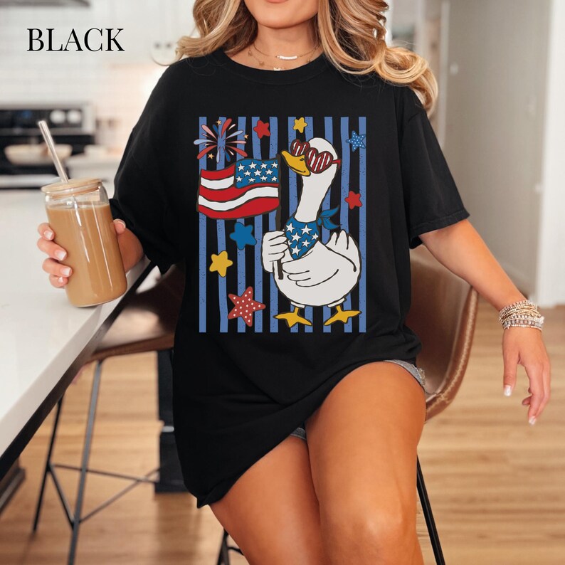 Pu&ograve; includere: T-shirt nero con un'oca dei cartoni animati con occhiali da sole e una bandana patriottica, che tiene una bandiera americana. Il design include stelle e strisce rosse, bianche e blu.