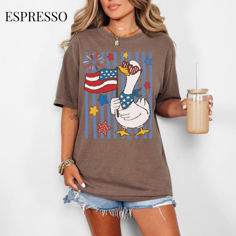 Pu&ograve; includere: T-shirt color espresso con un'oca dei cartoni animati con occhiali da sole e una bandana patriottica, che tiene in mano una bandiera americana. Il design include fuochi d'artificio, stelle e strisce blu. La parola "ESPRESSO" &egrave; stampata in alto a sinistra.