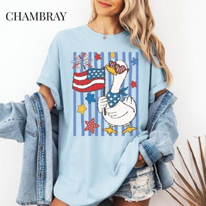 Pu&ograve; includere: T-shirt azzurra con un design patriottico. Presenta un'oca dei cartoni animati con occhiali da sole e una bandana, che tiene in mano una bandiera americana. La maglietta ha strisce blu verticali e accenti di stelle e fuochi d'artificio. La parola "CHAMBRAY" &egrave; stampata in alto.