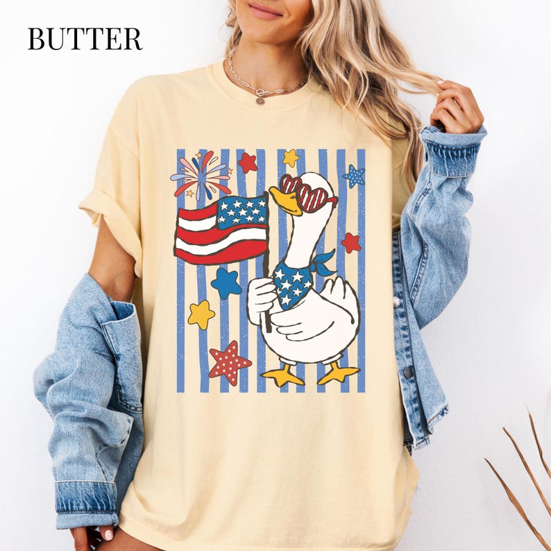 Pu&ograve; includere: T-shirt color burro con un'oca patriottica con occhiali da sole e una bandana, che tiene in mano una bandiera americana. Il design include stelle, fuochi d'artificio e strisce rosse, bianche e blu. La parola "BUTTER" &egrave; stampata in alto a sinistra.
