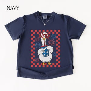 May include: Navy blue t-shirt featuring a cartoon goose wearing star-shaped glasses and holding a cup with stars and stripes. The goose is set against a red and white checkered background with fireworks.