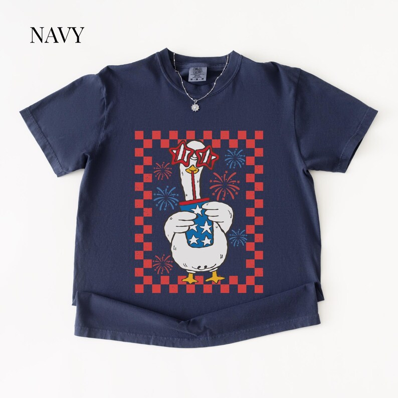 May include: Navy blue t-shirt featuring a cartoon goose wearing star-shaped glasses and holding a cup with stars and stripes. The goose is set against a red and white checkered background with fireworks.