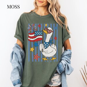 Pu&ograve; includere: T-shirt verde oliva con grafica di un'oca patriottica. L'oca indossa occhiali da sole e una bandana, con in mano una bandiera americana. Il design include stelle, fuochi d'artificio e strisce blu. La parola "MOSS" &egrave; nell'angolo in alto a sinistra.