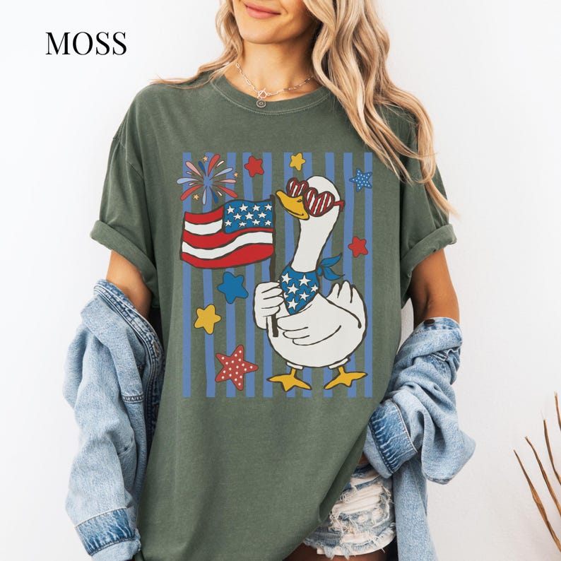 Pu&ograve; includere: T-shirt verde oliva con grafica di un'oca patriottica. L'oca indossa occhiali da sole e una bandana, con in mano una bandiera americana. Il design include stelle, fuochi d'artificio e strisce blu. La parola "MOSS" &egrave; nell'angolo in alto a sinistra.