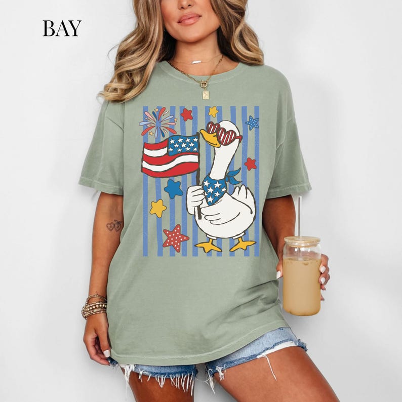Pu&ograve; includere: T-shirt verde salvia con un'oca dei cartoni animati con occhiali da sole e una bandana patriottica, che tiene in mano una bandiera americana. Il design include strisce blu, stelle e fuochi d'artificio. La parola "BAY" &egrave; in alto a sinistra.