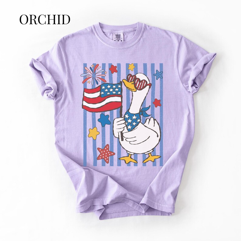 Pu&ograve; includere: Una maglietta lilla con un'oca dei cartoni animati con occhiali da sole e una bandana, che tiene una bandiera americana. Il disegno comprende stelle, fuochi d'artificio e strisce rosse, bianche e blu. La parola "ORCHID" &egrave; stampata in alto.