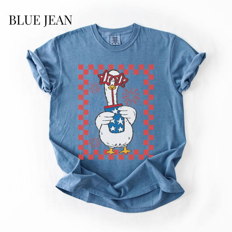 May include: Blue jean t-shirt featuring a cartoon goose wearing star-shaped glasses and holding a container with stars. The goose is framed by a red and white checkered pattern, with fireworks in the background. The text "BLUE JEAN" is at the top.