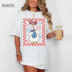 May include: White t-shirt featuring a cartoon goose wearing star-shaped glasses and holding a container with stars and stripes. The goose is set against a red and white checkered background with fireworks. The word "WHITE" is at the top left.