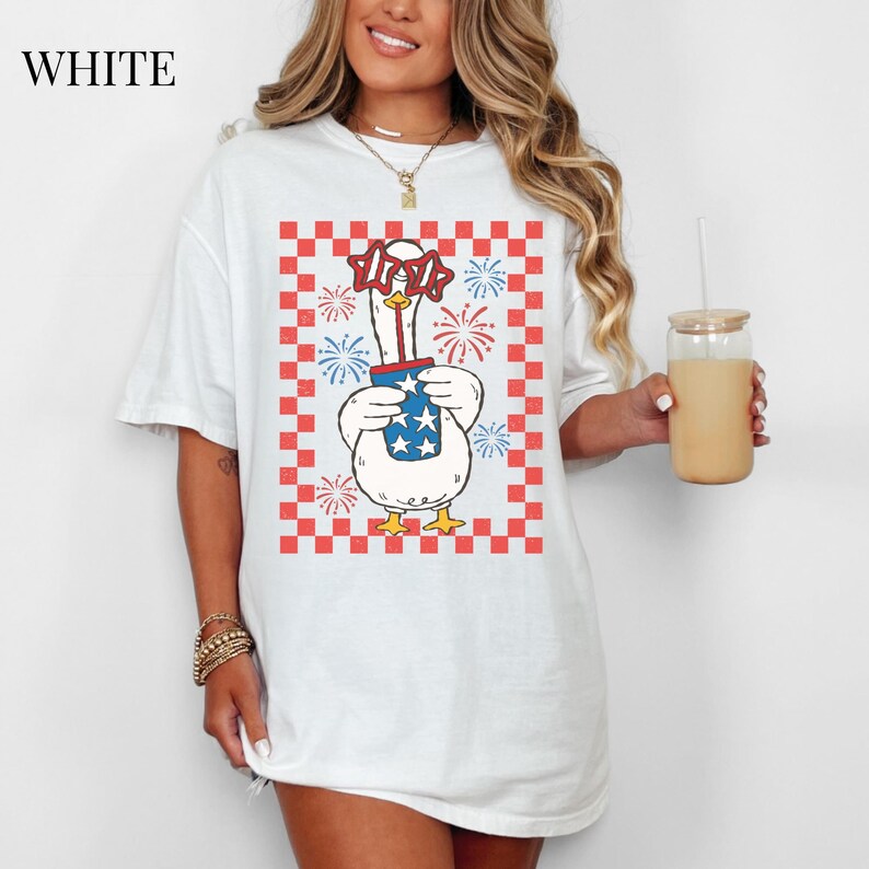 May include: White t-shirt featuring a cartoon goose wearing star-shaped glasses and holding a container with stars and stripes. The goose is set against a red and white checkered background with fireworks. The word "WHITE" is at the top left.