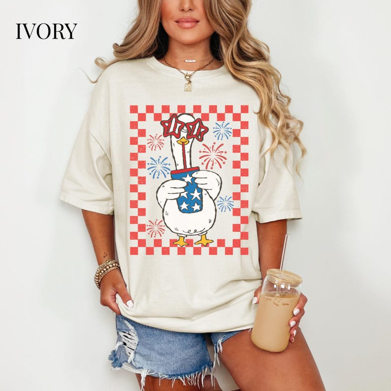 May include: Ivory-colored t-shirt featuring a cartoon goose wearing star-shaped glasses and holding a cup with stars. The goose is set against a red and white checkered background with fireworks. The word "IVORY" is printed at the top.