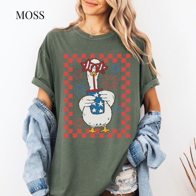 May include: Moss green t-shirt featuring a cartoon goose wearing star-shaped glasses and holding a cup with stars. The goose is set against a red and white checkered background with fireworks. The word "MOSS" is in the upper left corner.