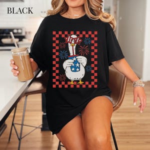 May include: Black t-shirt featuring a cartoon goose wearing patriotic glasses and holding a container with stars. The goose is framed by a red and black checkered pattern, with fireworks. The word "BLACK" is at the top.