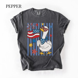 Pu&ograve; includere: T-shirt grigio scuro con un'oca dei cartoni animati con occhiali da sole e una bandana patriottica, che tiene una bandiera americana. Il design include stelle, fuochi d'artificio e la parola "PEPPER" in alto.