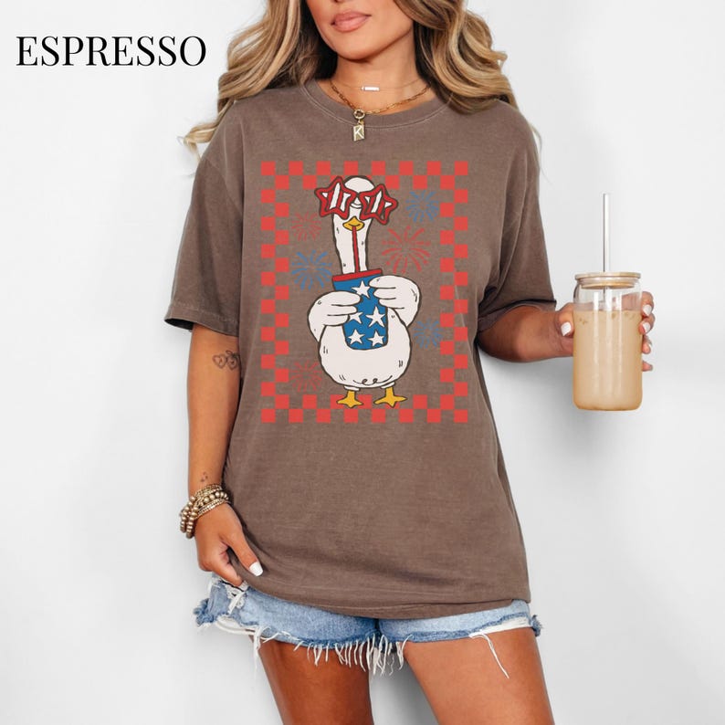 May include: Espresso-colored t-shirt featuring a cartoon goose wearing star-shaped glasses and holding a drink with stars. The goose is set against a red and white checkered background with fireworks. The word "USA" is on the glasses.