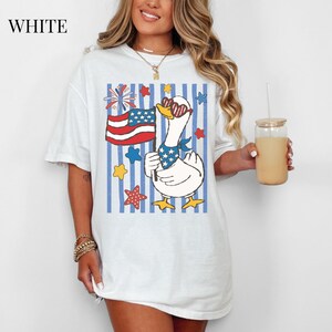 Pu&ograve; includere: T-shirt bianca con un'oca dei cartoni animati con occhiali da sole e una bandana patriottica, che tiene in mano una bandiera americana. Il design include stelle e strisce rosse, bianche e blu, con la scritta "WHITE" in alto a sinistra.
