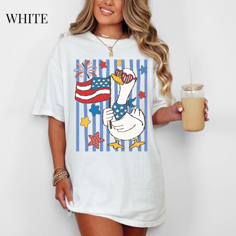 Pu&ograve; includere: T-shirt bianca con un'oca dei cartoni animati con occhiali da sole e una bandana patriottica, che tiene in mano una bandiera americana. Il design include stelle e strisce rosse, bianche e blu, con la scritta "WHITE" in alto a sinistra.