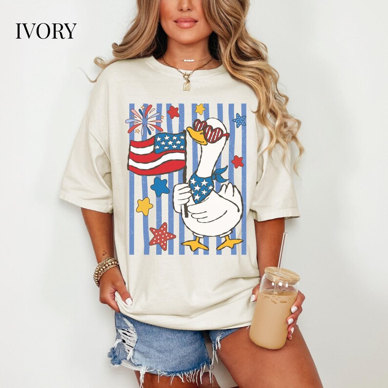 Pu&ograve; includere: T-shirt color avorio con un motivo di oca patriottica che indossa occhiali da sole e tiene in mano una bandiera americana. Il design include stelle e strisce rosse, bianche e blu, con la scritta "IVORY" in alto a sinistra.