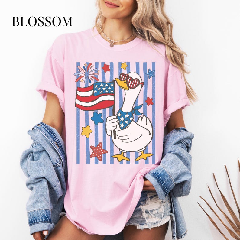Pu&ograve; includere: T-shirt rosa chiaro con grafica di un'oca patriottica. L'oca indossa occhiali da sole e una bandana a stelle, con una bandiera americana. Sfondo a righe blu e bianche con stelle e fuochi d'artificio. La parola "BLOSSOM" &egrave; stampata in alto.
